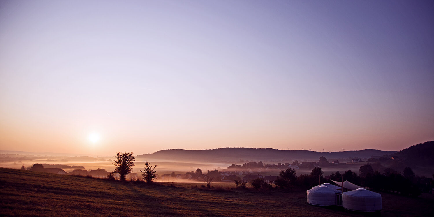 Jurten auf einer Wiese im Sonnenaufgang mit Blick auf Hügel und Nebel