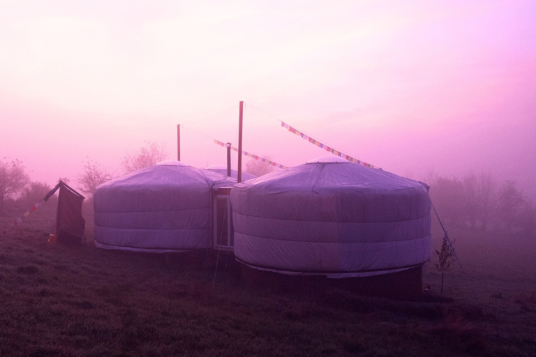 Miteinander verbundene Jurten mit Regentuch und Ofenrohr auf einer Wiese im dichten Morgennebel